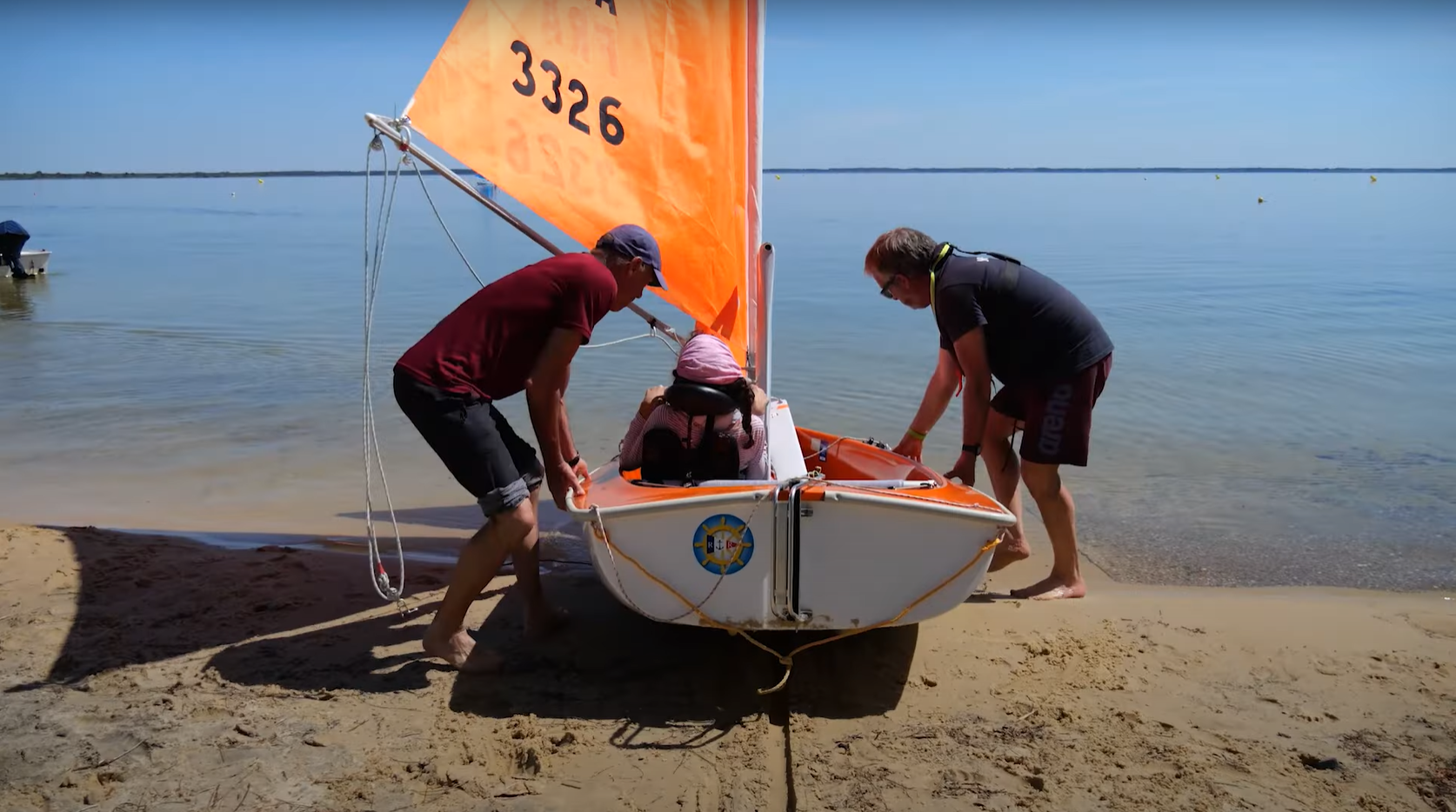 Deux bénévoles poussent un hansa depuis la plage jusqu'à l'eau. A son bord une pratiquante en situation de handicap physique prête à naviguer. 